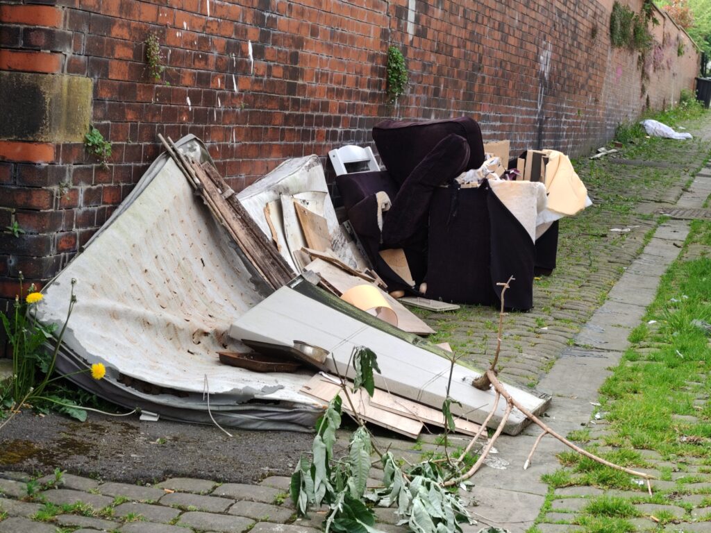 Photo of a back street with multiple items against a wall, including two dirty mattresses, broken wooden furniture and an old sofa