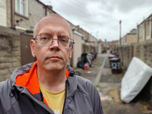 Martyn Hurt in the foreground, wearing a grey and orange jacket and yellow t-shirt. Out of focus in the background is a back street with a mattress on it on there right, wheelie bins in the fast background, and a glimpse of a black rubbish bag