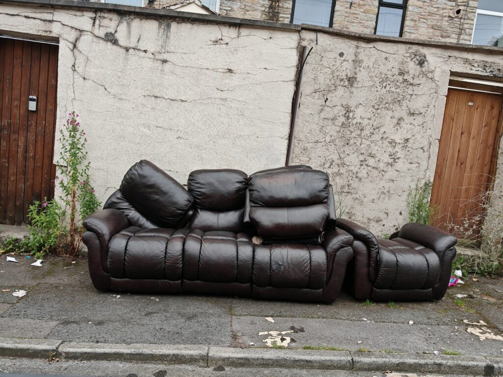 A brown leather sofa, recliner, and ottoman discarded on a residential street.