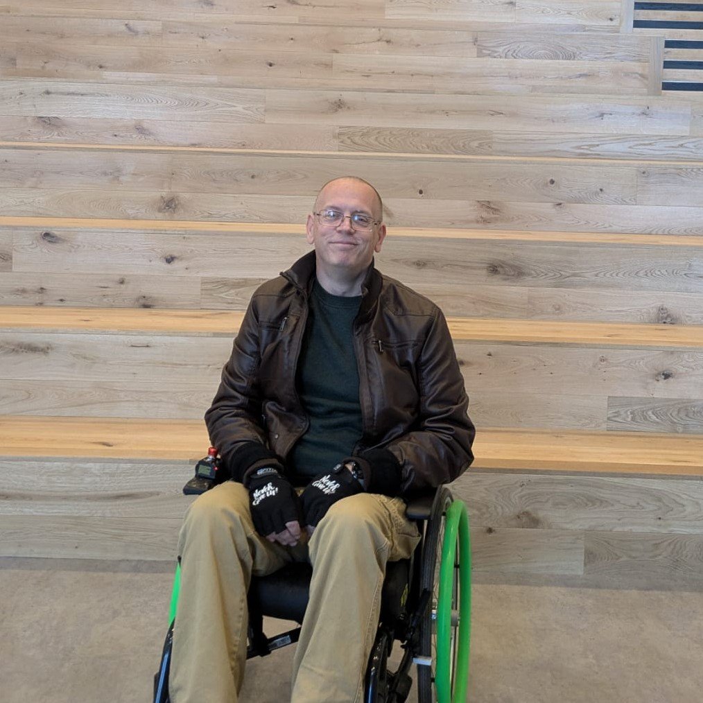 Martyn Hurt seated in a wheelchair with green pushrims on the wheels. He is wearing a dark brown leather jacket over a dark sweater, beige pants, and black gloves with white text on them. The man has glasses and a calm, content expression on his face. Behind him is a minimalist background featuring wooden tiered steps with a light natural finish. The setting appears modern and well-lit, suggesting an indoor location with a clean and contemporary design.