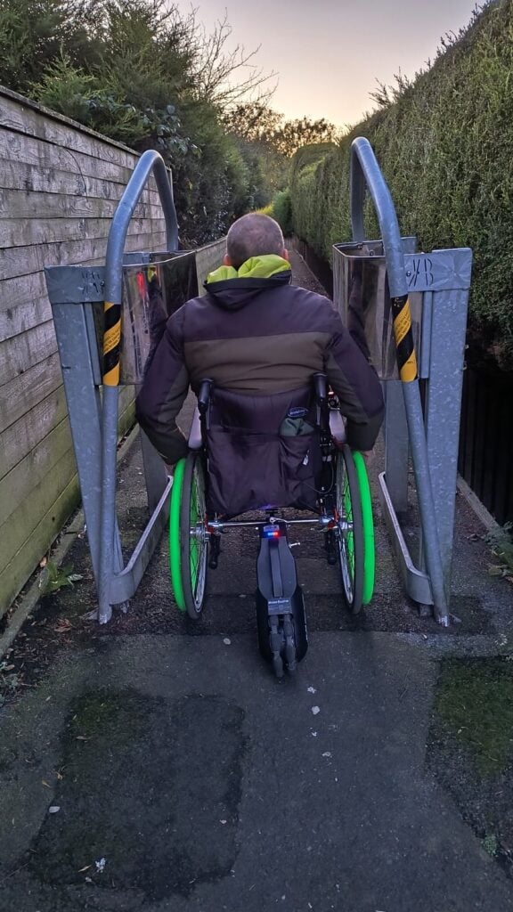 A rear view of a person in a wheelchair with bright green wheels navigating through a narrow metal gate along a pathway. The wheelchair has an attachment at the back, possibly a small motorized device. The individual is wearing a brown and green jacket with a hood. The pathway is bordered by a tall wooden fence on the left and dense green hedges on the right. The setting appears to be outdoors during early morning or late afternoon, with soft sunlight visible in the distance.
The scene highlights accessibility challenges in navigating tight spaces.